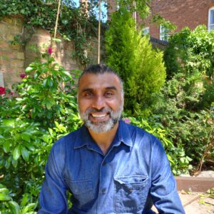 A smiling man in a blue shirt is sat in a garden on a bright day. 