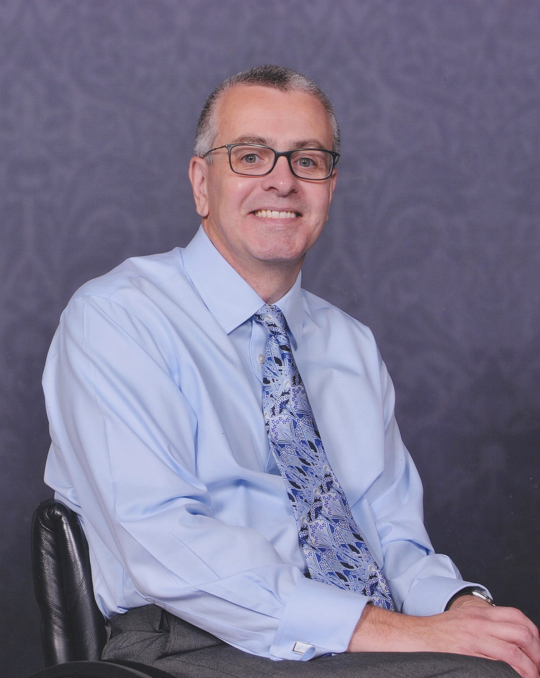 Portrait photo of Dr Tom Wells MBE. He is wearing a blue shirt, tie, glasses, and is smiling at the camera.
