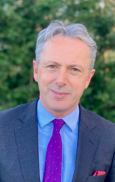 A profile photo of Judge Mark Sutherland Williams. He is smiling at the camera, and is wearing a grey suit with a purple tie.