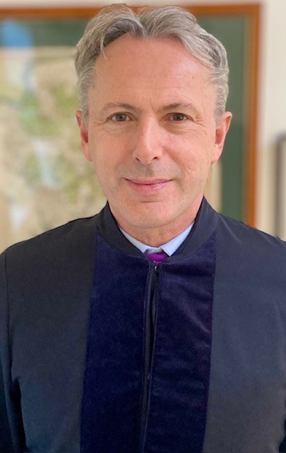 A profile photo of Judge Mark Sutherland Williams. He is smiling at the camera, and is wearing a blue suit.