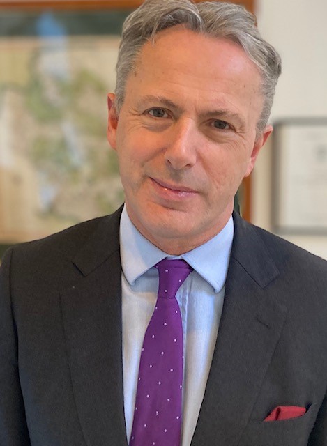 A profile photo of Judge Mark Sutherland Williams. He is smiling at the camera, and is wearing a grey suit with a purple tie.