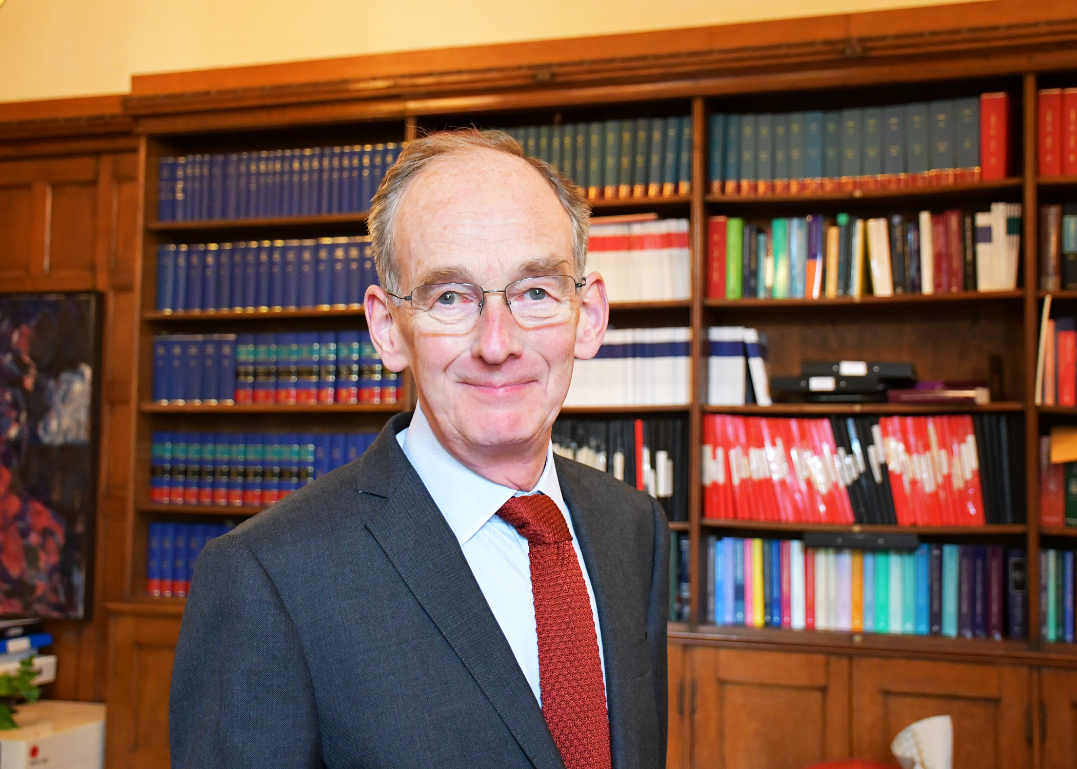 Sir Andrew McFarlane, smiling, in front of bookshelves, wearing a suit and tie